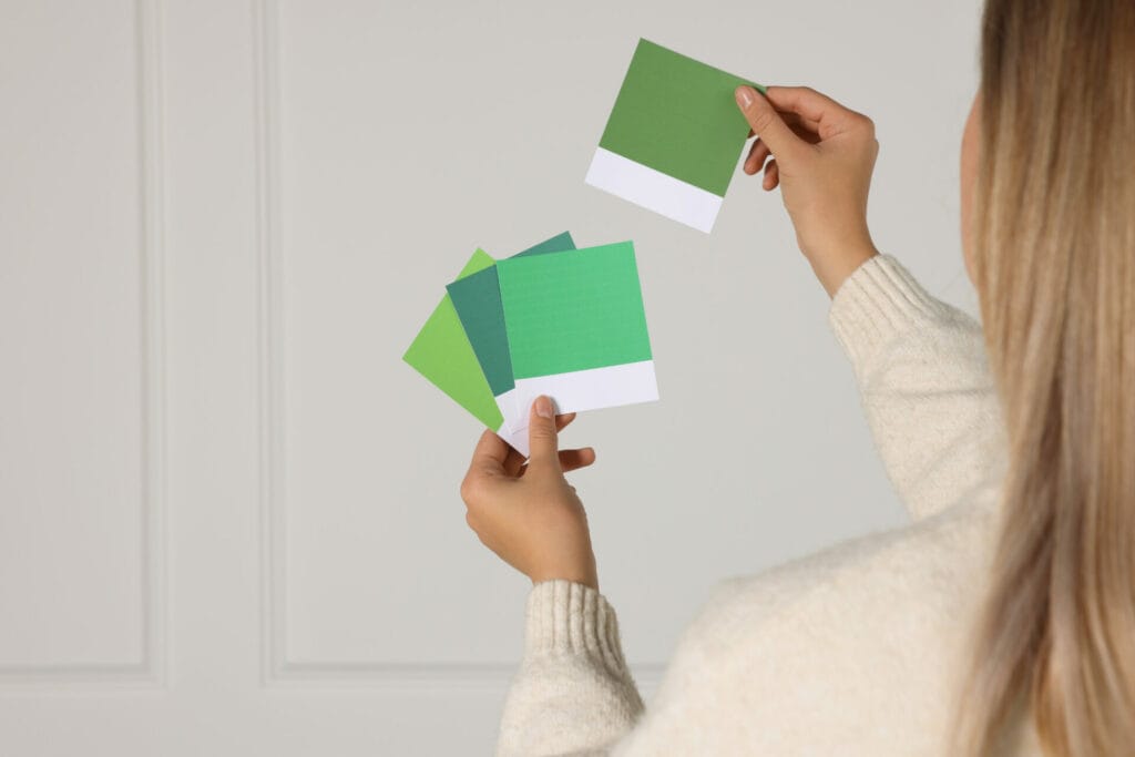 Woman choosing paint shade for wall indoors, focus on hands with color sample cards.