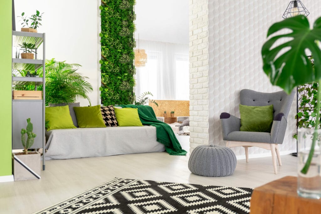 Sofa with green pillows and blanket standing in open space living room interior with grey armchair and footrest, black and white carpet and fresh plants
